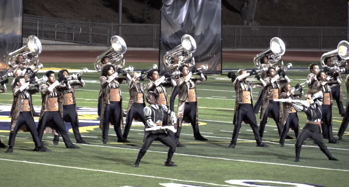 A marching band performs on at Cougar Stadium at College of the Canyons under stadium lights. The brass players, wearing matching ornate jackets and black pants, hold their instruments up in unison while two color guard members in contrasting silver and black outfits strike dynamic poses in front. The scene is highly synchronized and theatrical, with backdrops in the background and a dramatic sense of movement and energy.